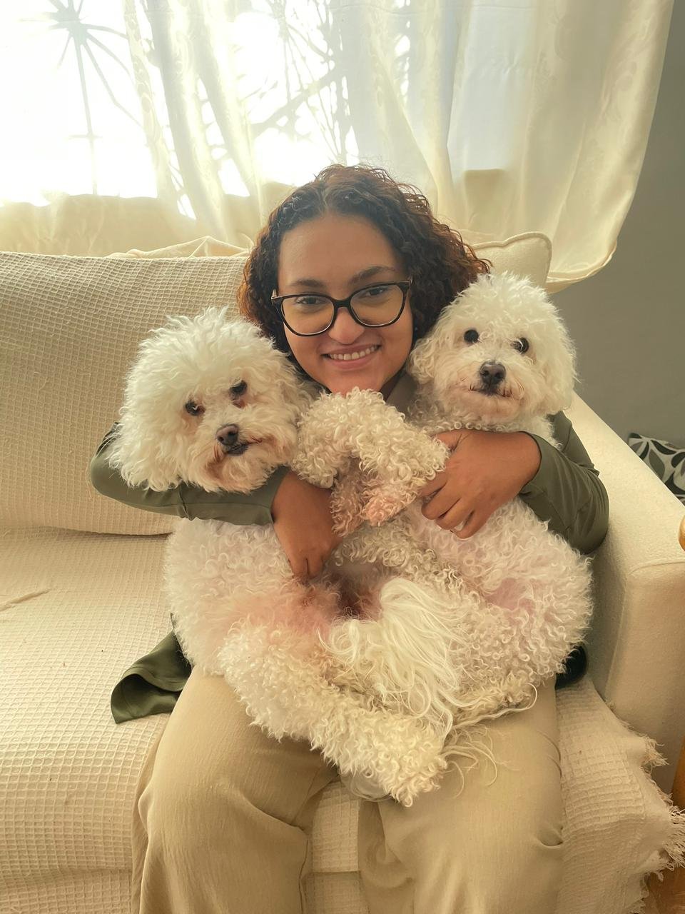 Dra. Mariana Helen sorrindo ao abraçar dois cachorrinhos brancos Bichon Frisé em atendimento domiciliar